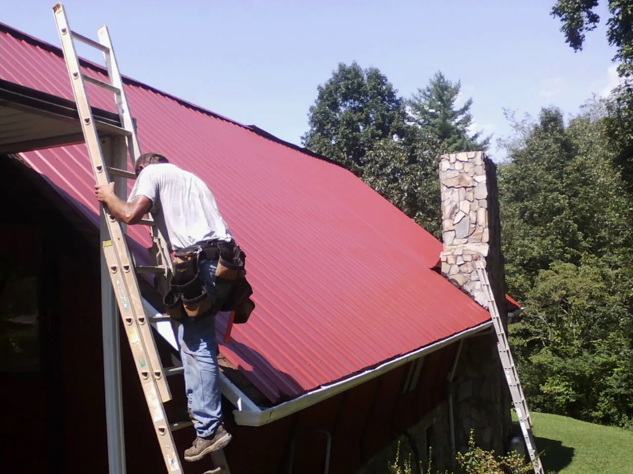 Roofer on a ladder beside a red metal roof and stone chimney — Soffit & Fascia Repair by Roofing Brothers in East Stroudsburg